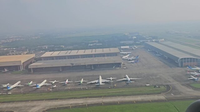 An expansive aerial view captures a commercial aircraft taking off from Soekarno-Hatta International Airport, revealing the massive GMF AeroAsia maintenance hangars, taxiways, and parked airplanes.