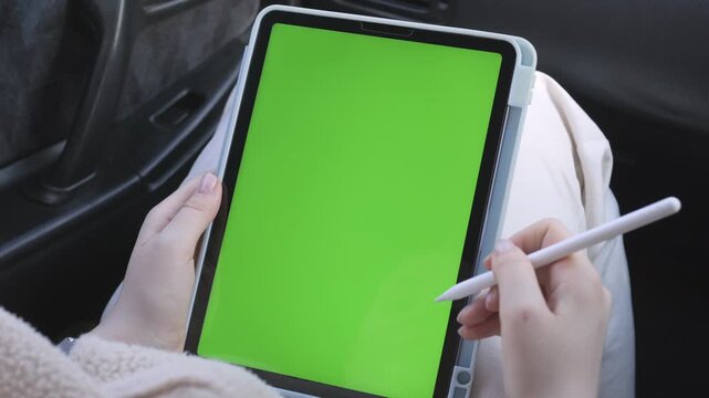 Person holds tablet with green screen in car. Hand uses stylus to draw or write on screen. Fingers steady as they interact with device. Car interior visible in background