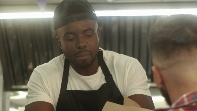 Close view of African American male handing food to customer. Man leaning across counter and giving orders. Wearing apron and cap. Working inside food truck kitchen during busy service moment.