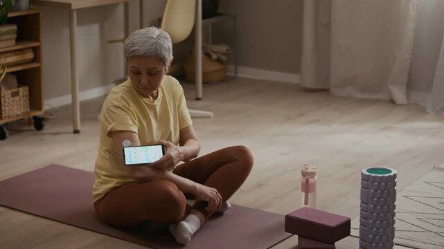 High angle shot of middle aged woman tracking blood sugar level on smartphone app connected to CGM device on upper arm during home fitness session