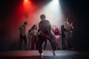 Fototapeta premium Young male dancer posing under red and white stage lights with group behind. Concept of dance school ad, music video casting, performer portfolio, club event and theater promo.