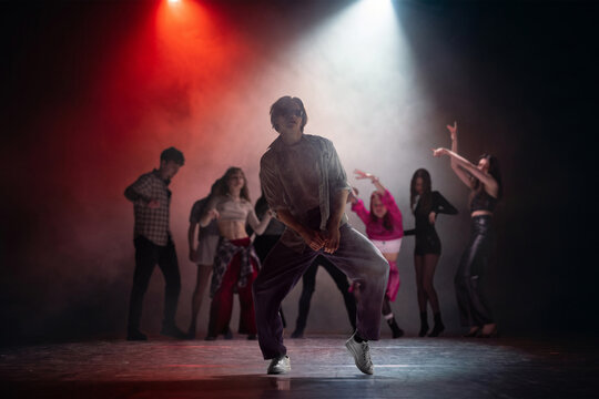 Young male dancer posing under red and white stage lights with group behind. Concept of dance school ad, music video casting, performer portfolio, club event and theater promo.