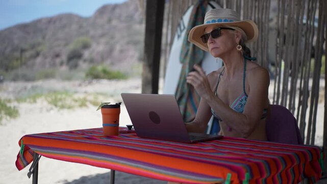Mature woman works on her computer at a tropical beach while on vacation, showing emotion with sunlight shining around her.