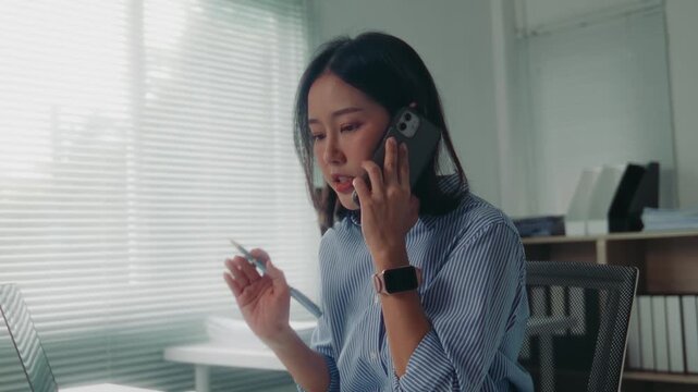 Anxious asian businesswoman feeling stressed and frustrated while talking on her mobile phone and working on her laptop at the office