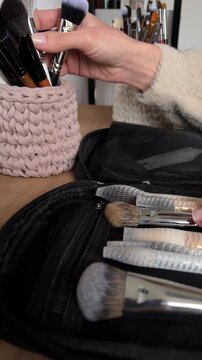 Close up angled view of female hands transferring professional makeup brushes from a black zippered organizer with mesh protectors into a pink knitted brush holder on a wooden desk, demonstrating beau
