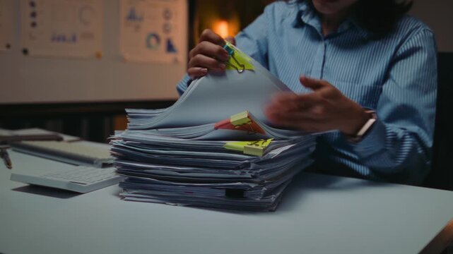 Businesswoman sorting through a large stack of paperwork with colorful sticky notes at her desk. Hardworking employee working overtime in a dark office