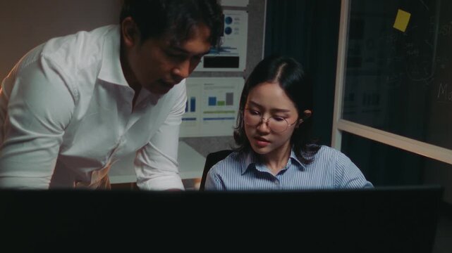 Two business people working on a project late at night. The man, likely a manager, is helping his female colleague with her work on the computer