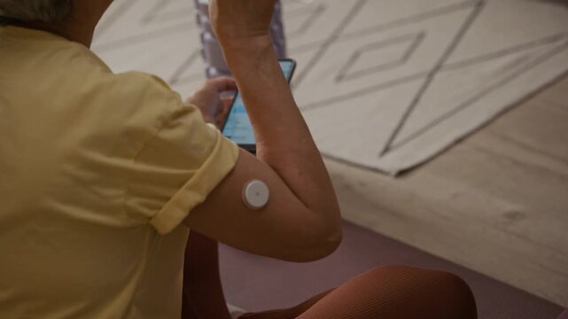 Over shoulder shot of middle aged woman drinking water while monitoring blood sugar readings on mobile app connected wearable CGM sensor during home yoga session