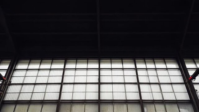 Empty industrial warehouse interior foregrounding a vast wall of grimy windows, revealing the interplay of light and shadow on the decaying industrial architecture