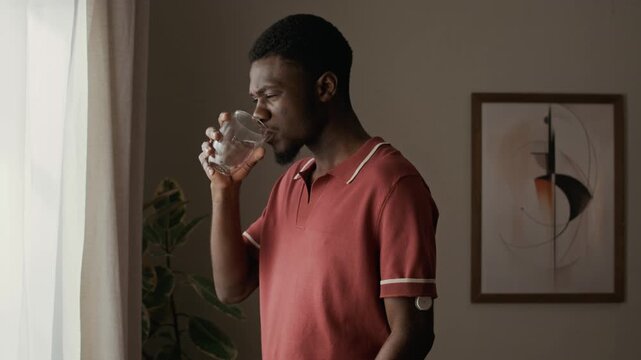 Medium shot of young adult Black man with diabetes wearing continuous glucose monitor on arm drinking water beside window in living room