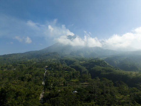 Aerial view of Mount Merapi volcano surrounded by lush green forests and clouds under a clear blue sky in Kecamatan Cangkringan, Daerah Istimewa Yogya, Indonesia.