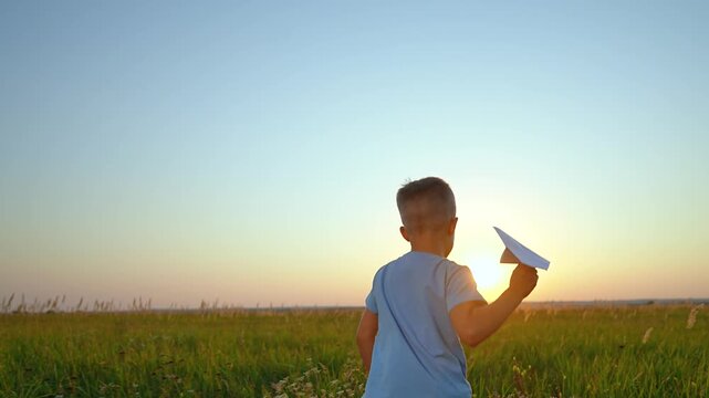 Boy launches paper airplane in field at sunset. Child throws paper plane outdoors. Kid plays with aircraft in meadow. Boy launching glider at golden hour. Child throws airplane in field.