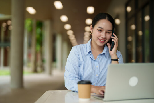 Young Asian businesswoman multitasking, having a phone call while typing on a laptop and enjoying coffee outdoors, representing a modern mobile office or distant work lifestyle