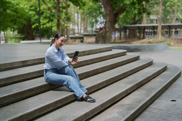 Fototapeta premium Woman sitting on campus steps using a mobile phone, engaging with digital content, representing modern communication, remote work, and online education