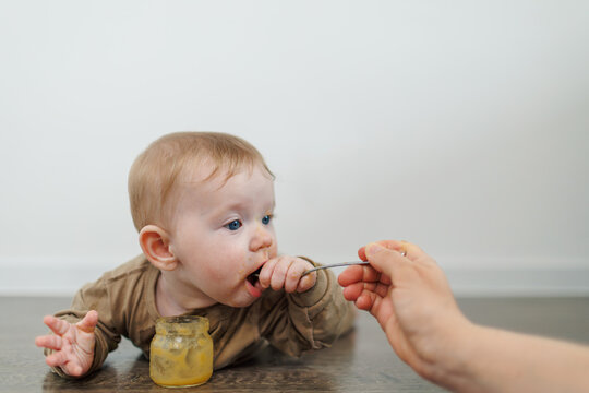 Infant lying on the floor is being spoon-fed. The baby looks to the side while tasting the food. A small glass jar of puree stands on the dark wooden surface