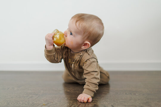 A baby sitting on the floor tries to eat directly from a small glass jar of puree. The child's clothes are heavily stained with food