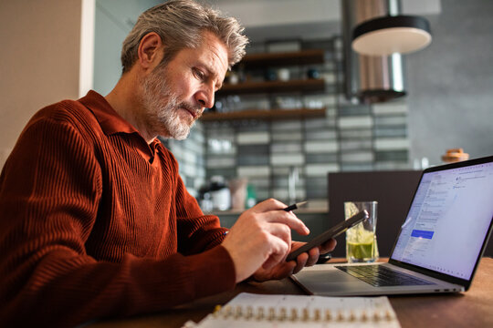 Mature man working from home with laptop and smartphone in modern kitchen