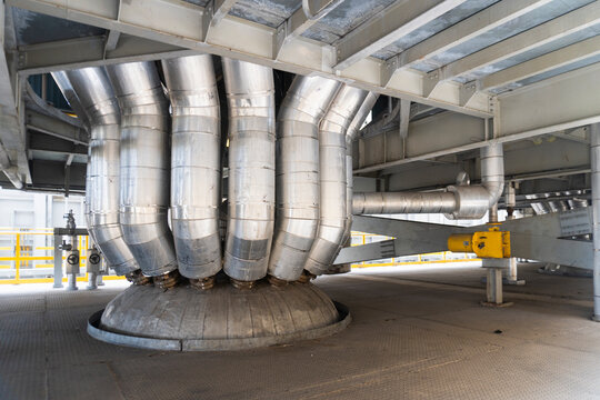 Bottom view of a massive industrial burner system with silver insulated pipes connected to a central unit in a power plant