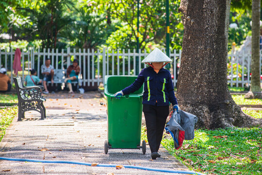 A sanitation worker wearing a traditional Vietnamese conical hat (Non La) pushing a green trash bin in a sunny park in Ho Chi Minh City
