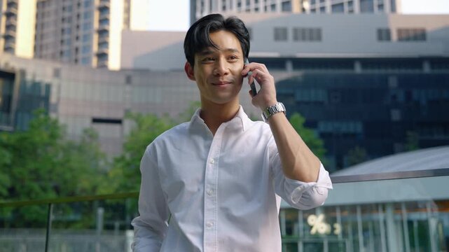Confident young Asian businessman making a phone call outside modern office buildings. Corporate communication and professional business lifestyle in urban environment.
