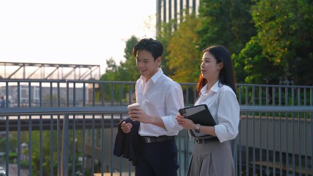 Young Asian businessman and businesswoman walking together in an urban office area during golden hour, discussing work while holding coffee cup and tablet device. Modern corporate lifestyle and teamwo