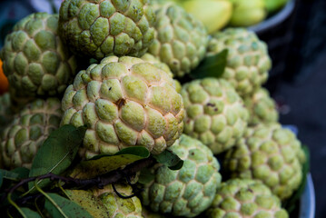 Fototapeta premium Green, bumpy custard apples piled together. Fruits fresh, with leaves still attached. Market stall display. Perfect for food blogs, tropical recipes, or healthy eating content
