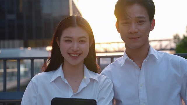 Portrait of young Asian businessman and businesswoman smiling confidently at camera in modern business district during sunset. Corporate teamwork and professional success concept.
