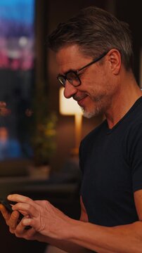Smiling adult man using cell phone in a cozy evening indoor setting at home, with city lights in the background.