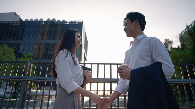 Young Asian business professionals having conversation outside office building while holding coffee cup and tablet device. Modern teamwork and collaboration in urban workplace.
