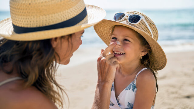 Mother applying sunscreen to daughter's nose at the beach, sun protection skincare routine, day at seaside, family bonding under sun, summer vacation fun, sun safety awareness, outdoor leisure