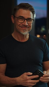 Casual mid adult man with cell phone at home. Home office, banking online, checking social media. Portrait of older bearded guy smiling. Using mobile application, telecommunication.
