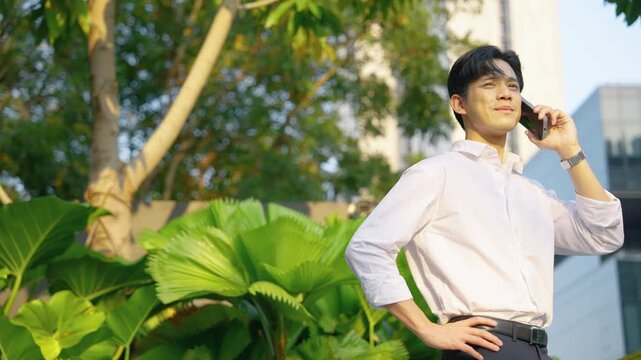 Young Asian businessman speaking on smartphone while standing outdoors near modern office buildings and green plants. Professional communication and corporate lifestyle concept.
