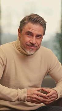 Portrait of confident middle aged man sitting at home, wearing turtleneck pullover. Mid adult older male in cosy living room, happy, smiling.