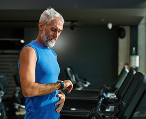 Fototapeta premium Portrait of a senior man exercising in a gym, mature male running using treadmill machine and checking smart watch for performance, healthy lifestyle and cardio exercise at fitness club concepts, vita
