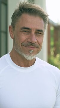 Portrait of relaxed middle-aged man smiling enjoying garden on sunny day. Happy handsome male in 50s with gray hair and beard on terrace at home.