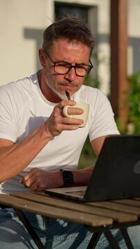 Businessman working remotely in garden. Mature man using laptop outdoor drinking morning coffee. Male entrepreneur freelancer tele working online in home office. Digital business lifestyle.