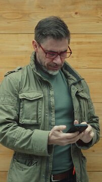 Bearded man wearing glasses and green jacket using smartphone against a wooden wall. Mature mid adult, middle aged male smiling with phone. Telecommunication and connectivity.