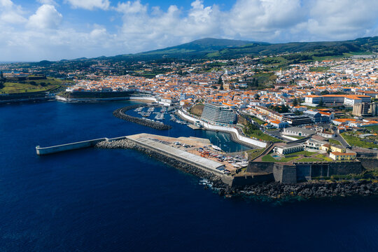 Aerial view of the historic coastal city featuring white buildings with terracotta roofs, a harbor with breakwaters, and lush green hills under a blue sky Angra do Heroismo, Azores, Portugal.