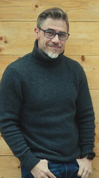 Portrait of middle aged man outdoor in front of wooden wall wearing pullover. Happy satisfied older male smiling. Eyeglasses and gray hair. Autumn or spring clothes.