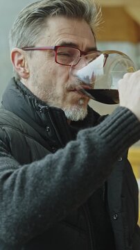 Expert drinking, tasting glass of red wine. Middle aged man outdoor on home balcony in wooden cabin wearing turtleneck pullover and warm vest. Eyeglasses and gray hair. Autumn or winter.