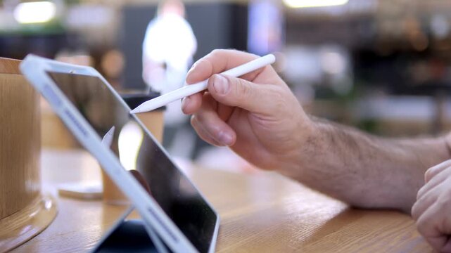 Hand holds stylus over tablet on wooden table. Coffee cup beside device. Background blurred with warm lighting. Person focused on digital work. Casual indoor setting, likely cafe or workspace