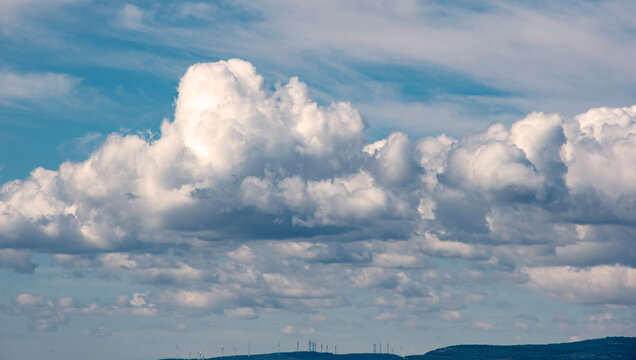 Cielo azul con grandes nubes blancas tipo c&uacute;mulo sobre colinas lejanas donde se ven aerogeneradores, creando una escena natural y tecnol&oacute;gica armoniosa.