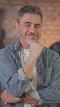 Portrait of happy casual middle aged man standing at home arms crossed in loft living room. Senior male with grey hair in denim shirt looking at camera, smiling, thinking.
