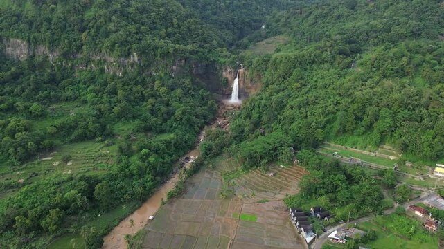 Drone aerial view of a majestic waterfall, named Curug Cimarinjung, in a lush tropical jungle, surrounded by beautiful green rice terrace, river, and small rural village, located in WestJava Indonesia