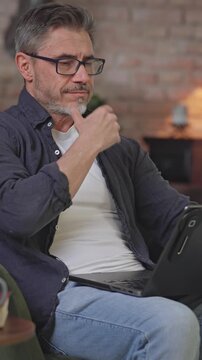 Serious confident businessman working with laptop at home office desk. Middle aged man in glasses sitting in armchair, concentrating, browsing internet. Male entrepreneur remote managing business.