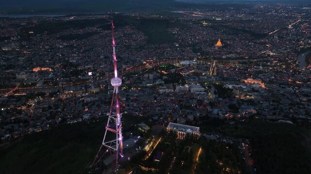 Aerial view of Tbilisi TV Tower illuminated at night with city lights and Holy Trinity Cathedral in the background, Georgia cityscape from Mtatsminda Park at dusk.