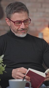Smiling middle aged man sitting in living room reading book. Senior male wearing turtleneck pullover and eyeglasses. Smart looking older guy with grey hair enjoying leisure and free time at home.