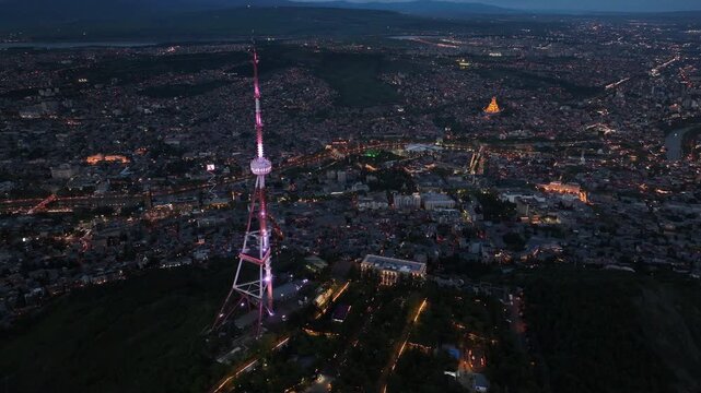 Aerial view of Tbilisi TV Tower illuminated at night with city lights and Holy Trinity Cathedral in the background, Georgia cityscape from Mtatsminda Park at dusk.