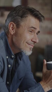Confident older white man at home, sitting on couch in dark living room, relaxing, drinking coffee or tea. Portrait of happy, mature age, middle age, mid adult caucasian man in 50s, smiling.