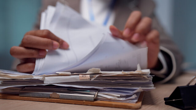 Hands sort through thick stacks of paper documents. Fingers gently flip pages with focused attention. Desk holds binders and folders beneath the hands. Background shows soft blue tones